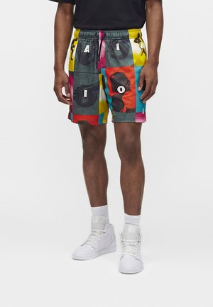 Homme portant un short coloré à motifs avec des lettres et des graphismes, des baskets montantes blanches et des chaussettes blanches, debout devant un fond uni.