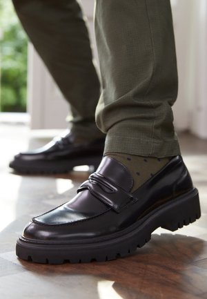 Schwarze Lederloafer mit dicken Sohlen, getragen zu olivgrünen Hosen und gemusterten Socken auf einem Holzboden in der Nähe einer hellen Türöffnung.