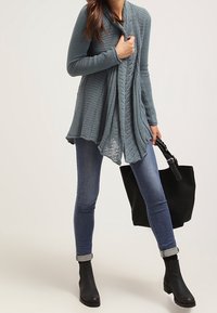 Petrolblå stickad cardigan med draperad front, kombinerad med åtsittande jeans och svarta ankelstövlar. En svart handväska fulländar outfiten.