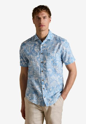 Mann trägt ein hellblaues kurzärmeliges Hemd mit weißen tropischen Drucken und beige Hose, steht mit einer Hand in der Tasche.