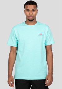 Camiseta de manga corta turquesa en algodón ligero, con un pequeño parche rectangular de la marca en el lado izquierdo del pecho con texto rojo y azul.