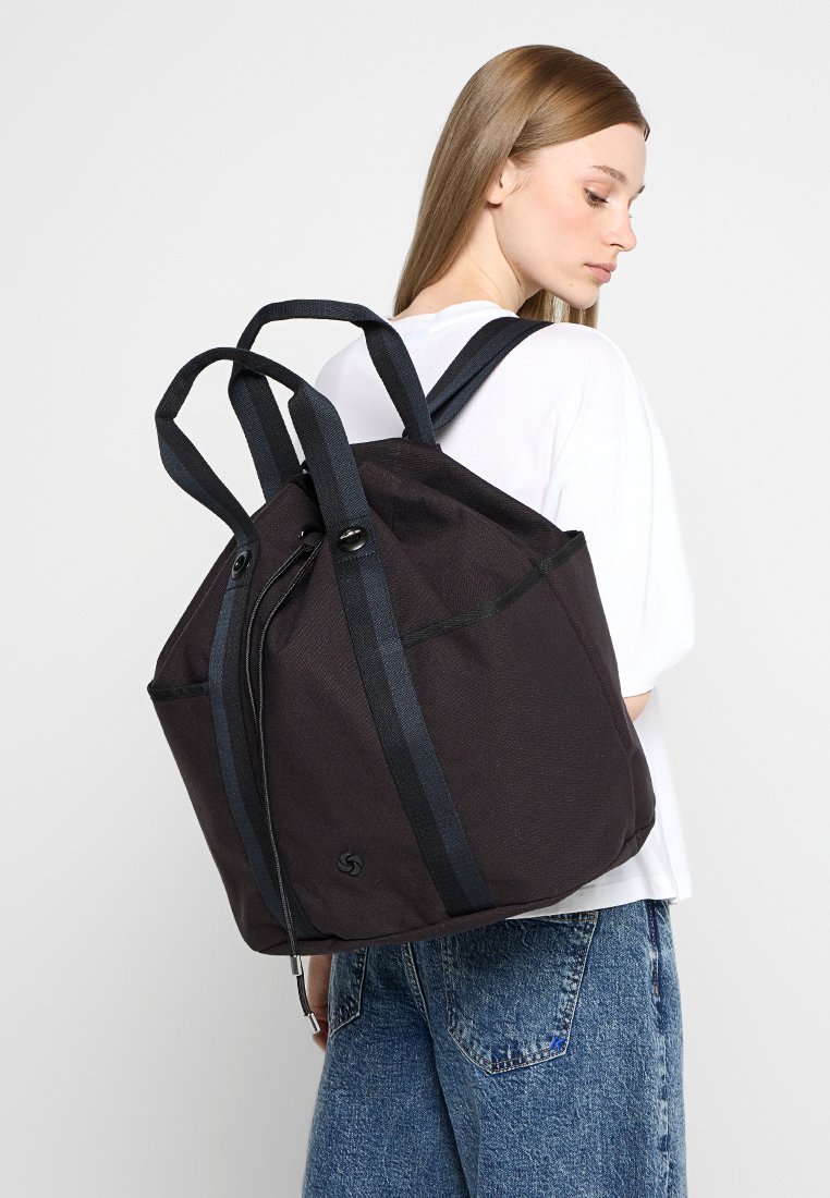 Mochila de tela negra con múltiples correas ajustables, una superficie texturizada y un cierre con cremallera en la parte superior. Presenta un detalle de logo sutil.