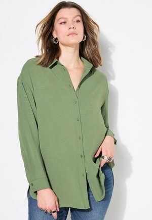 Grøn langærmet skjorte med knaplukning, krave, blød tekstur, sideslids og afslappet pasform, parret med blå jeans og ringe.