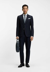 Marynarka granatowa na dwa guziki, biała koszula wizytowa, wzorzysty krawat oraz czarne skórzane buty. Trzyma ciemnoniebieską skórzaną teczkę.