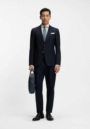 Marineblauer, maßgeschneiderter Anzug mit zwei Knöpfen, weißes Hemd, gemusterte Krawatte und schwarze Lederschuhe. Hält eine dunkelblaue Lederaktentasche.