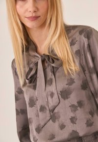 Blusa gris con cuello atado, textura suave y un sutil patrón floral en tonos más oscuros. Mangas largas con un ajuste holgado.