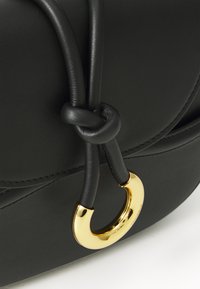 Borsa in pelle nera con una texture liscia, dotata di una maniglia annodata e di un accento circolare dorato. Design curvo con dettagli cuciti.