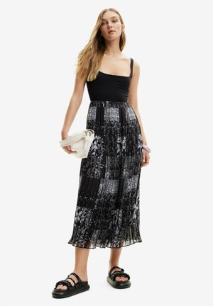 Débardeur noir associé à une jupe maxi plissée à motif floral en argent et noir. Le modèle tient une pochette blanche. Sandales noires à glissière.