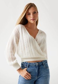 Młoda kobieta z długimi blond włosami nosi biały sweter dzianinowy z dekoltem w serek oraz niebieskie dżinsy z wysokim stanem, pozując z jedną ręką w kieszeni.