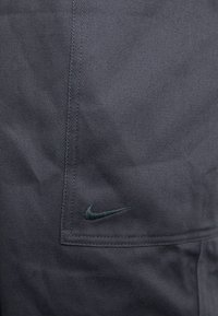 Tamno sive hlače od tkanine s teksturiranim završetkom, detaljnim šavovima i malim tamnozelenim Nike logotipom na bočnoj džepu.