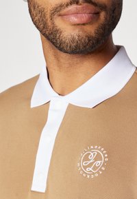 Beige polo krekls ar baltu apkakli, ar teksturētu audumu un izšūtu logotipu kreisajā krūtī, ietver baltas pogu aizdares.