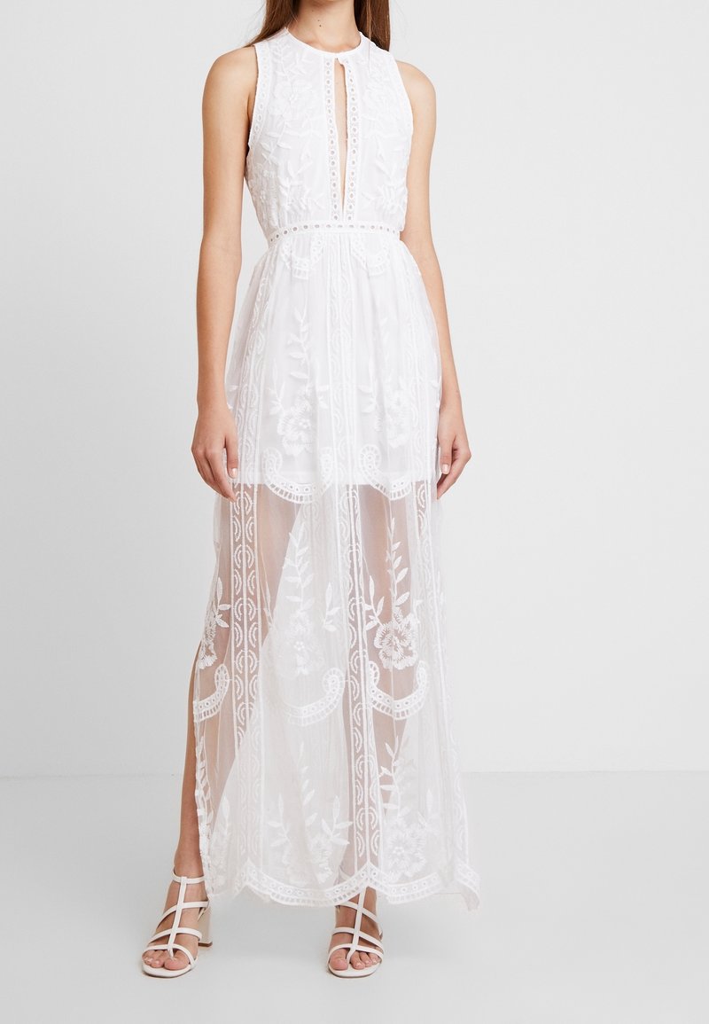 Femme portant une robe maxi en dentelle blanche sans manches avec broderie florale et une jupe transparente sur une doublure plus courte, associée à des talons blancs à brides.
