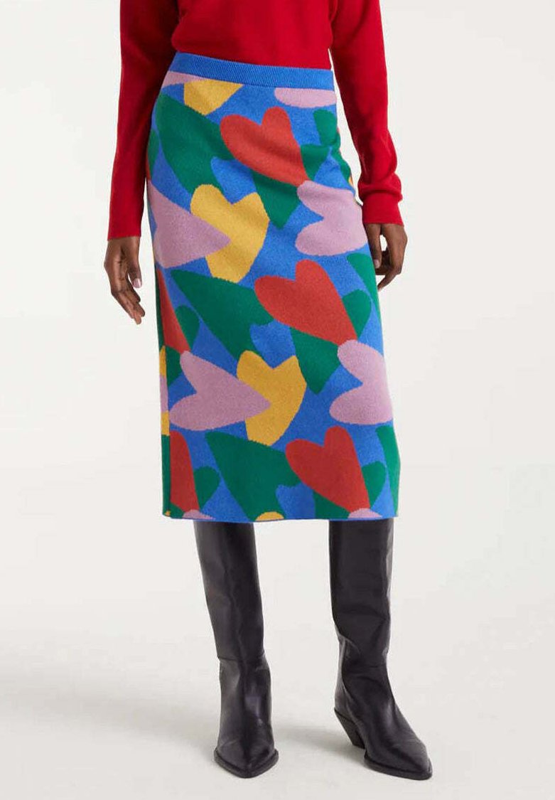 Falda hasta la rodilla con formas de corazones superpuestos en rojo, verde, amarillo y rosa sobre un fondo azul, combinada con botas negras y una blusa roja.
