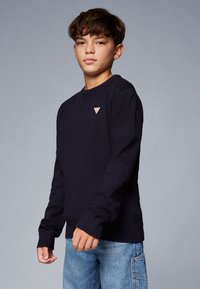 Sweat-shirt en tricot bleu marine à manches longues, avec des poignets et un ourlet côtelés, arborant un petit logo en forme de triangle sur la poitrine. Porté avec un jean cargo bleu.