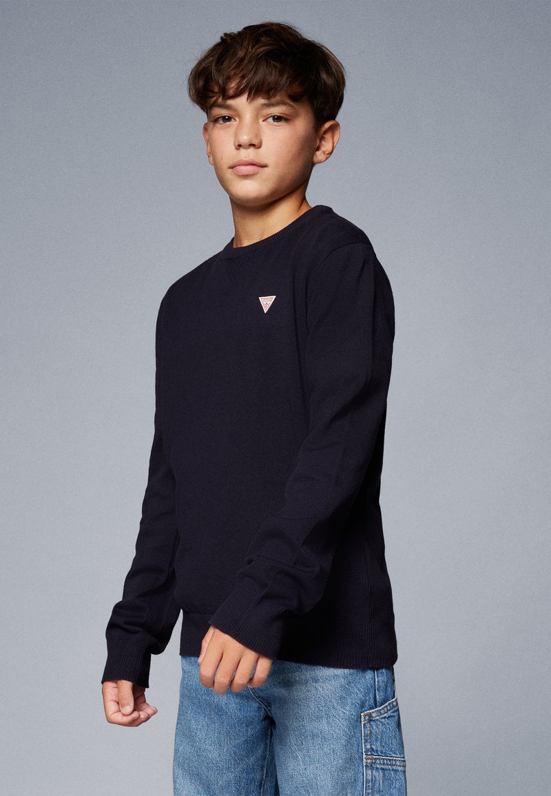 Sweat-shirt en tricot bleu marine à manches longues, avec des poignets et un ourlet côtelés, arborant un petit logo en forme de triangle sur la poitrine. Porté avec un jean cargo bleu.