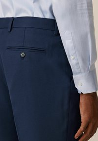 Pantalones de vestir en color navy con una tela texturizada, un bolsillo trasero y un detalle de botón. Combinados con una camisa azul clara con puños abotonados.