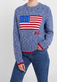Pull en tricot bleu avec un grand motif de drapeau américain, accents rouges sur les poignets et l'ourlet, et coupe décontractée. Idéal pour un style décontracté.