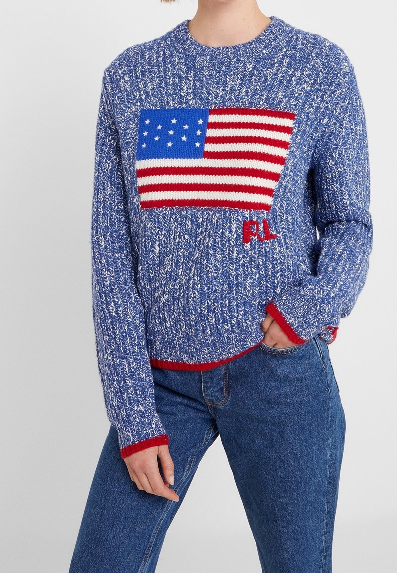 Pull en tricot bleu avec un grand motif de drapeau américain, accents rouges sur les poignets et l'ourlet, et coupe décontractée. Idéal pour un style décontracté.