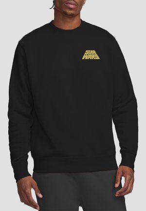 Man wearing a black sweatshirt with a small yellow "Star Wars" logo on the left chest standing against a plain light background.