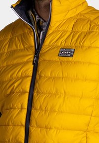 Geel gewatteerde jas met een geborduurde textuur, voorzak met rits sluiting en een marineblauwe binnenkant. Heeft een merkpatch op het borstgebied.