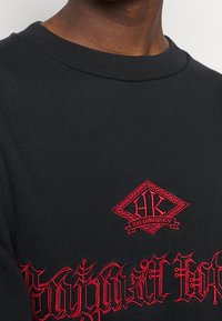 Svart t-shirt med röd broderad logga på bröstet och dekorativ text. Rund halsringning med ribbad textur.