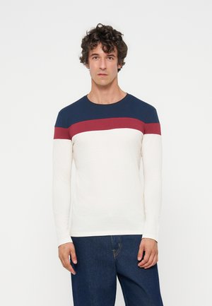 Homme aux cheveux bouclés portant une chemise à manches longues avec des blocs de couleur horizontaux bleu marine, rouge et blanc et un jean bleu foncé, debout, face à l’avant.
