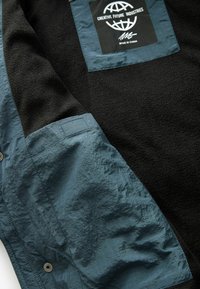 Veste en polaire avec un corps noir, des accents en tissu synthétique bleu clair et un patch logo ; elle présente une texture douce et une fermeture par bouton.