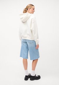 Hoodie crema con cappuccio, vestibilità rilassata, abbinato a pantaloni corti di jeans blu chiaro con orli sfilacciati. Scarpe platform nere e calze bianche.
