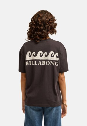 Personne aux cheveux bruns bouclés portant un t-shirt Billabong noir avec des motifs de vagues blanches, associée à un jean bleu, de dos.