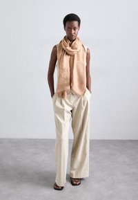 Sciarpa beige con un motivo sottile drappeggiata attorno al collo, indossata sopra un top senza maniche e pantaloni cream a pieghe e morbidi. Sandali completano l'look.