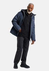 Giacca impermeabile blu con cappuccio, indossata sopra un pile nero con zip. Pantaloni e scarpe da trekking neri. Focus su funzionalità e comfort.