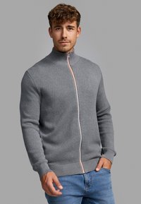 Grå zip-up genser med et strukturert mønster, høy krage og kontrasterende oransje glidelåsd tilbehør. Brukt med blå jeans.
