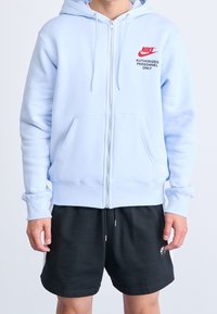 Sudadera con cremallera en azul claro, hecha de una tela suave, que cuenta con un bolsillo frontal, puños acanalados y un logotipo de Nike en rojo con texto negro en el pecho.