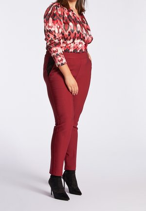 Vrouw die een rood en roze blouse met lange mouwen met patroon draagt, rode aansluitende broek en zwarte puntige hoge hakken enkellaarzen, staand opzij.
