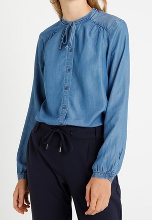 Femme portant un chemisier bleu à manches longues boutonné avec un détail noué au col et un pantalon noir avec cordon de serrage.
