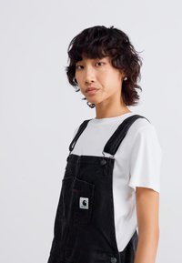 Schwarze Jeanslatzhosen mit einer Fronttasche, verstellbaren Trägern und einem kleinen Logopatch, getragen über einem weißen Rundhals-T-Shirt. Lockige Haare.