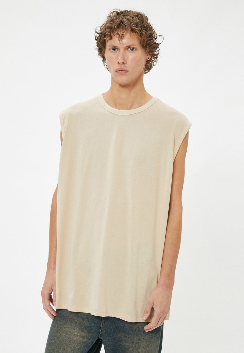 Beige mouwloze T-shirt gemaakt van zachte stof, met een ronde halslijn en een relaxte pasvorm, met zichtbare stiksels langs de zoom en randen.