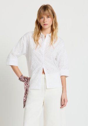 Blonde Frau trägt eine weiße Lochstickerei-Bluse und weiße Hose, hält einen roten Paisley-Schal in der linken Hand vor einem schlichten Hintergrund.