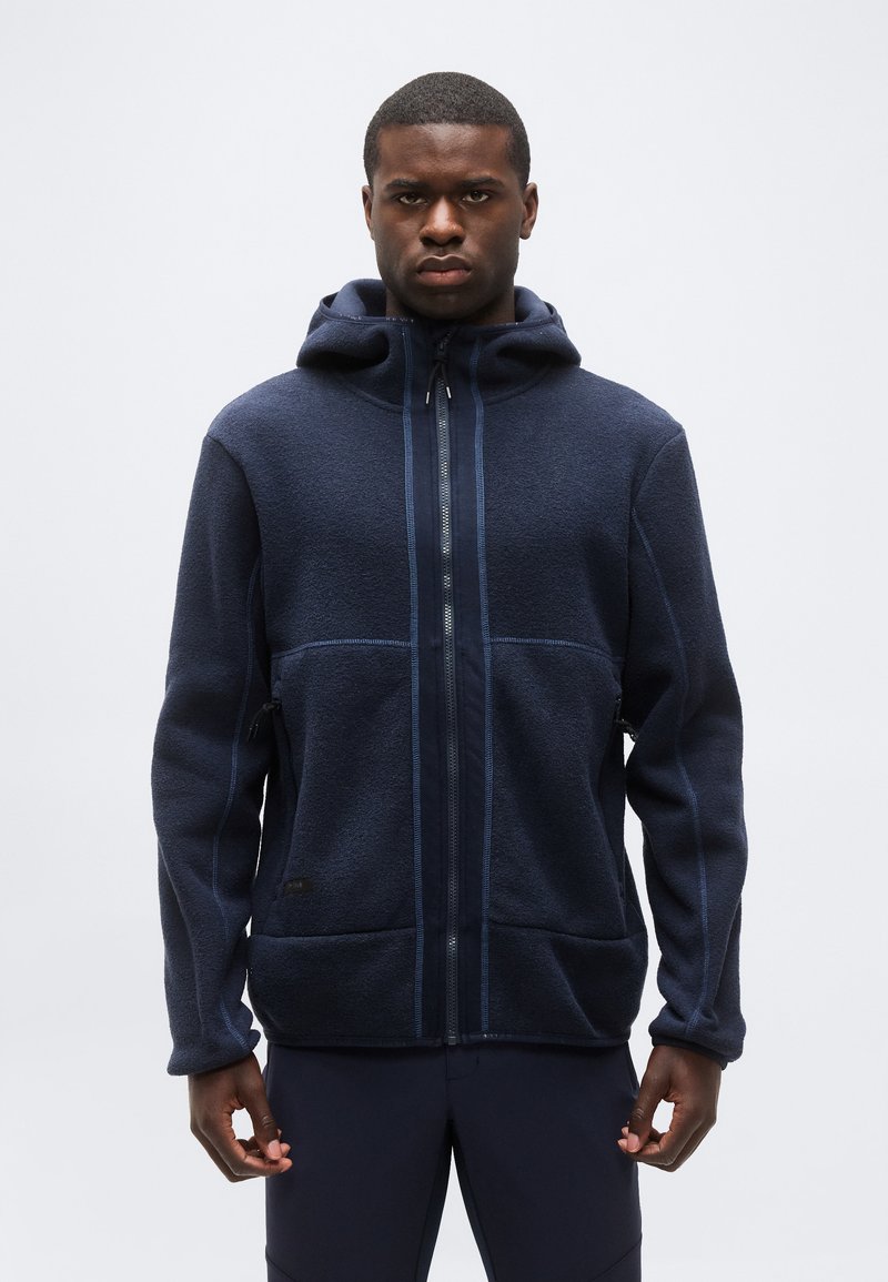 Νέος άνδρας που φοράει ζακέτα φλις με κουκούλα σε ναυτικό μπλε χρώμα, στέκεται μπροστά σε απλό ανοιχτό γκρι φόντο.