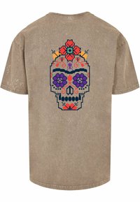 Beige t-skjorte med kort erm som har et fargerikt pikslet hodeskalle-design på baksiden med blomsterdetaljer i rødt, lilla og blått.