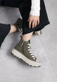 Zapatillas altas de color verde oliva con cordones blancos y una cremallera decorativa. Presentan una suela blanca gruesa con detalles texturizados.