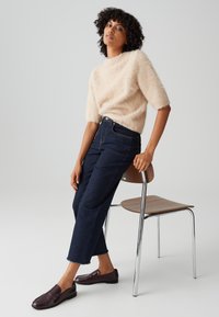 Beige fuzzy sweater with short sleeves, dark blue high-waisted jeans, and burgundy loafers, seated on a wooden chair with chrome legs.