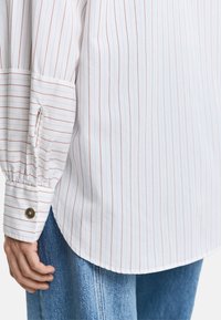 Hvit skjorte med smale røde striper, lange ermer, knappesperre-detaljer og buet hem. Kombinert med blå denim.