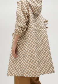 Person iført en beige og hvid ternet frakke med hætte og rynkede ærmer, stående med armene afslappet ned langs siderne.