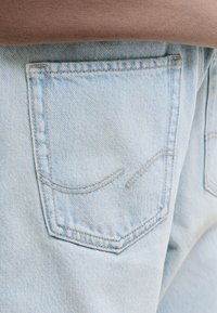 Τσέπη από ανοιχτό μπλε τζιν με μπεζ ραφές, φορεμένη με καφέ τοπ, που δείχνει μέρος της πλάτης και των πλευρικών ραφών.