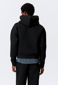 Sort hættetrøje med afslappet pasform, blød tekstur og ribbede ærmegab, lagdelt over en blå skjorte, stylet med matchende sorte bukser.