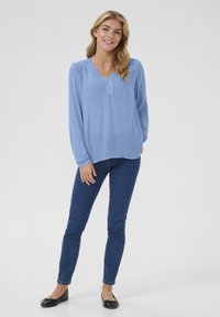 Blouse à manches longues bleu clair avec un décolleté en V, associée à un jean skinny en denim foncé et des chaussures plates noires. Texture de tissu lisse.