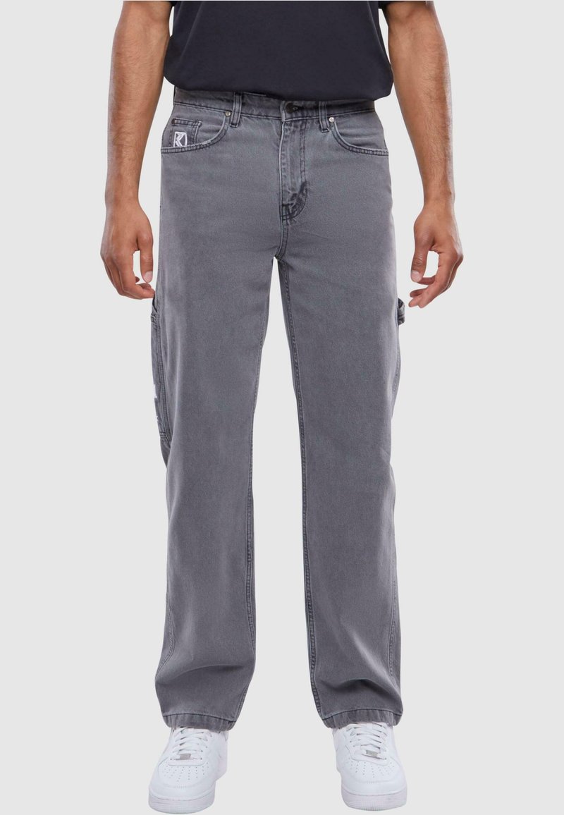 Pantalones cargo grises con un corte relajado, que cuentan con bolsillos laterales y un lavado desgastado. Combinados con zapatillas blancas y una camiseta negra.