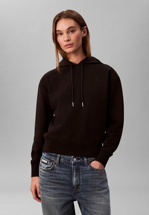 Ung kvinde iført en sort hættetrøje og blå jeans, stående med den ene hånd i lommen foran en ensfarvet grå baggrund.