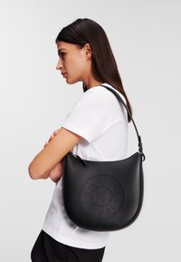 Sac à épaule en cuir noir de forme arrondie, avec un design de logo perforé. Bandoulière ajustable et texture lisse.